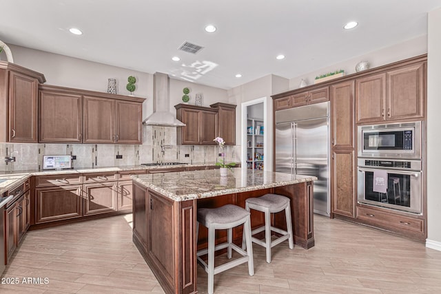 kitchen with light stone countertops, decorative backsplash, wall chimney exhaust hood, built in appliances, and a kitchen island