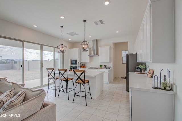 kitchen with wall chimney exhaust hood, an island with sink, white cabinetry, appliances with stainless steel finishes, and decorative light fixtures