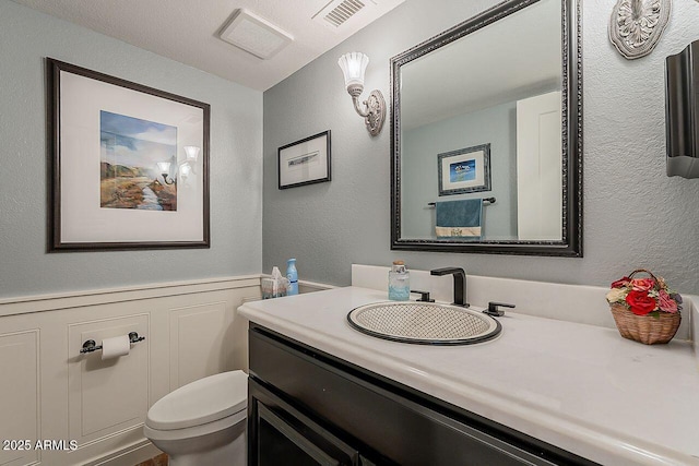 bathroom featuring a wainscoted wall, visible vents, a textured wall, toilet, and vanity