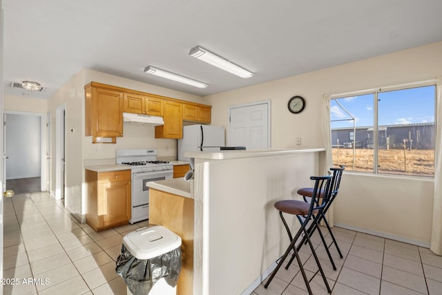 kitchen with light tile patterned floors, a breakfast bar area, under cabinet range hood, white appliances, and light countertops