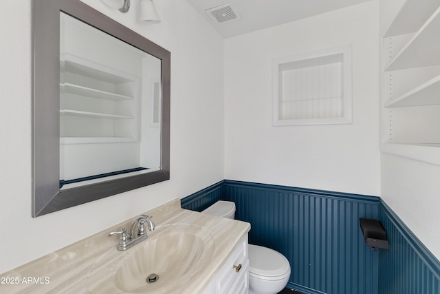 half bath with vanity, toilet, visible vents, and a wainscoted wall