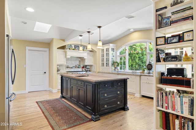 kitchen featuring lofted ceiling with skylight, an island with sink, white cabinets, stainless steel fridge, and light hardwood / wood-style flooring