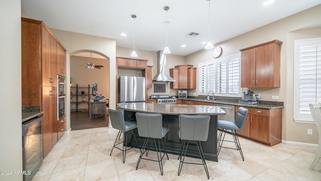 kitchen with a center island, wall chimney range hood, ceiling fan, appliances with stainless steel finishes, and decorative light fixtures