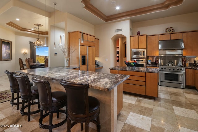 kitchen featuring a tray ceiling, built in appliances, decorative light fixtures, and dark stone countertops