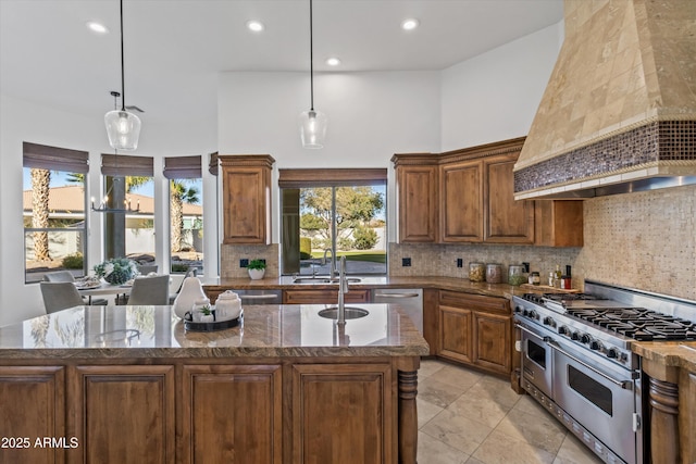 kitchen with premium range hood, hanging light fixtures, a high ceiling, stainless steel appliances, and a center island with sink