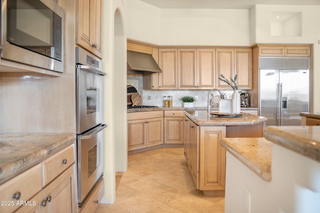 kitchen with wall chimney exhaust hood, light stone countertops, stainless steel appliances, light brown cabinets, and backsplash