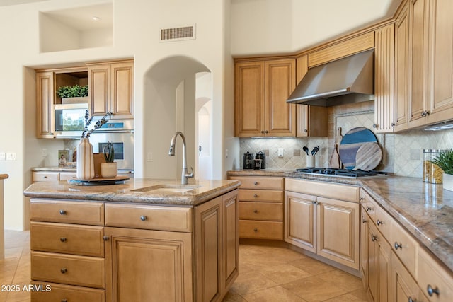 kitchen featuring arched walkways, appliances with stainless steel finishes, a kitchen island with sink, a sink, and wall chimney exhaust hood