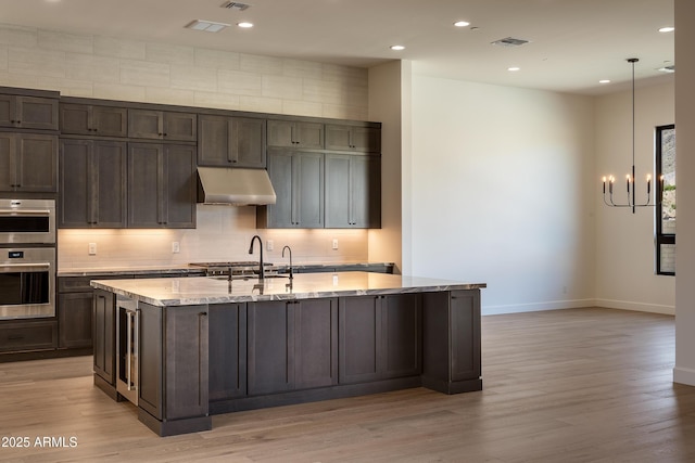 kitchen featuring backsplash, light stone countertops, under cabinet range hood, an island with sink, and a notable chandelier