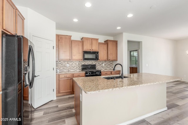 kitchen featuring light wood finished floors, backsplash, a sink, an island with sink, and black appliances