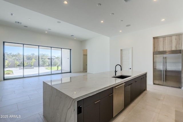 kitchen with sink, appliances with stainless steel finishes, a kitchen island with sink, dark brown cabinets, and light stone countertops