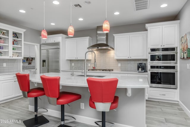 kitchen featuring a kitchen bar, wall chimney exhaust hood, an island with sink, stainless steel appliances, and white cabinets