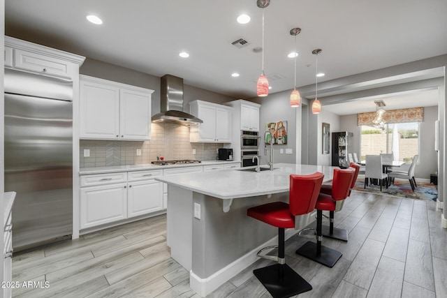 kitchen with an island with sink, appliances with stainless steel finishes, wall chimney range hood, and white cabinets