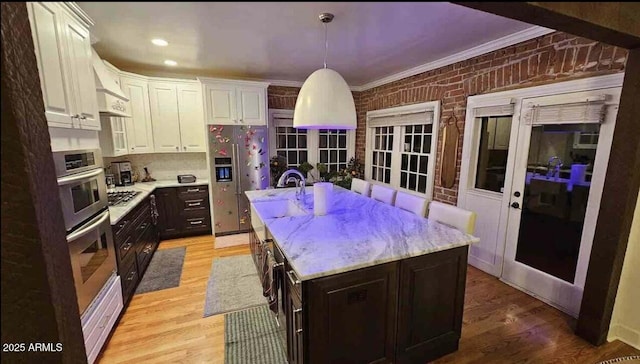 kitchen featuring a center island with sink, hanging light fixtures, appliances with stainless steel finishes, white cabinetry, and brick wall