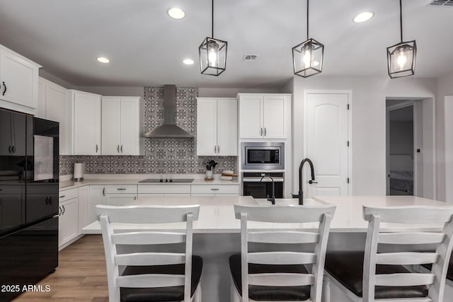 kitchen with appliances with stainless steel finishes, white cabinetry, an island with sink, decorative light fixtures, and wall chimney exhaust hood