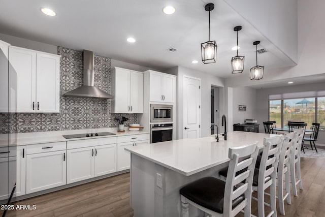 kitchen featuring wall chimney exhaust hood, stainless steel appliances, a center island with sink, and white cabinets