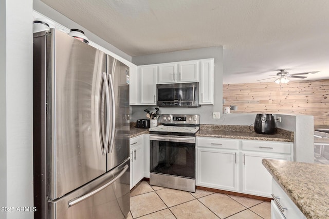 kitchen with light tile patterned flooring, wood walls, appliances with stainless steel finishes, ceiling fan, and white cabinets