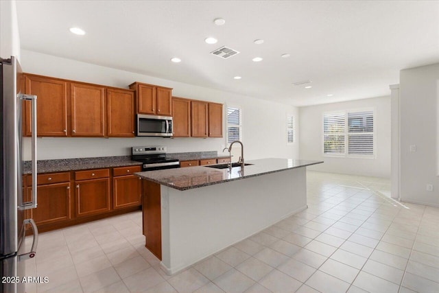 kitchen featuring sink, light tile patterned flooring, dark stone countertops, an island with sink, and appliances with stainless steel finishes