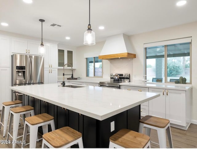 kitchen featuring a center island with sink, custom range hood, appliances with stainless steel finishes, decorative light fixtures, and white cabinetry