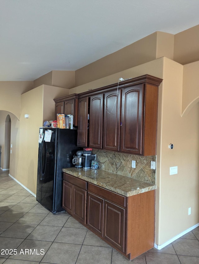 kitchen with dark brown cabinetry, light stone counters, light tile patterned floors, black refrigerator, and backsplash