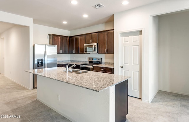 kitchen featuring stainless steel appliances, a kitchen island with sink, a sink, dark brown cabinetry, and light stone countertops