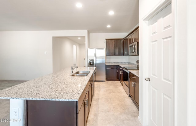 kitchen with dark brown cabinetry, light stone countertops, a kitchen island with sink, stainless steel appliances, and a sink