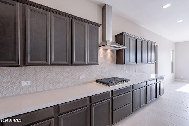 kitchen with stainless steel gas stovetop, backsplash, wall chimney range hood, light tile patterned floors, and dark brown cabinetry