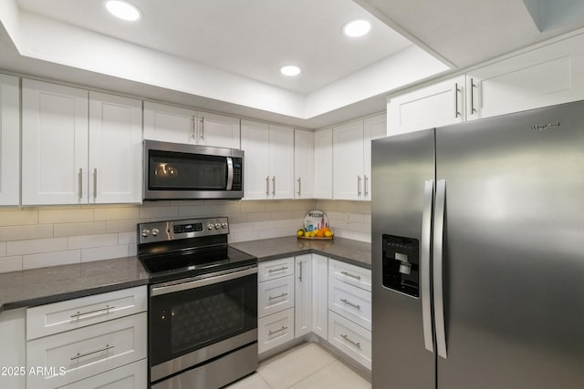 kitchen with appliances with stainless steel finishes, a tray ceiling, white cabinetry, decorative backsplash, and light tile patterned flooring