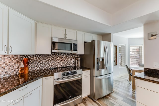 kitchen with stainless steel appliances, tasteful backsplash, white cabinetry, light wood-type flooring, and dark stone counters