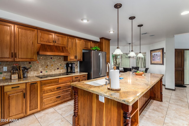 kitchen with hanging light fixtures, a center island with sink, stainless steel refrigerator, custom range hood, and black electric stovetop