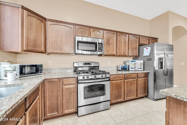 kitchen with appliances with stainless steel finishes, brown cabinetry, light tile patterned flooring, and light stone countertops