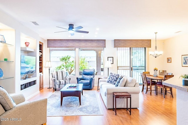 living room with built in shelves, a healthy amount of sunlight, ceiling fan with notable chandelier, and light hardwood / wood-style floors