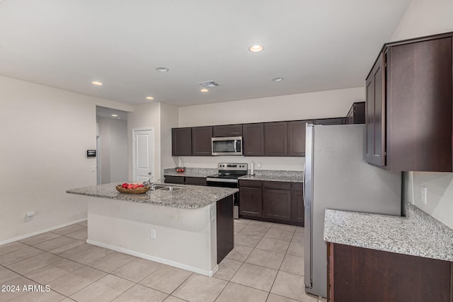 kitchen featuring a center island with sink, light tile patterned floors, appliances with stainless steel finishes, light stone countertops, and dark brown cabinetry