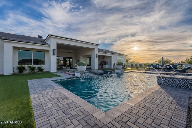 pool at dusk with pool water feature, a patio area, a lawn, and ceiling fan