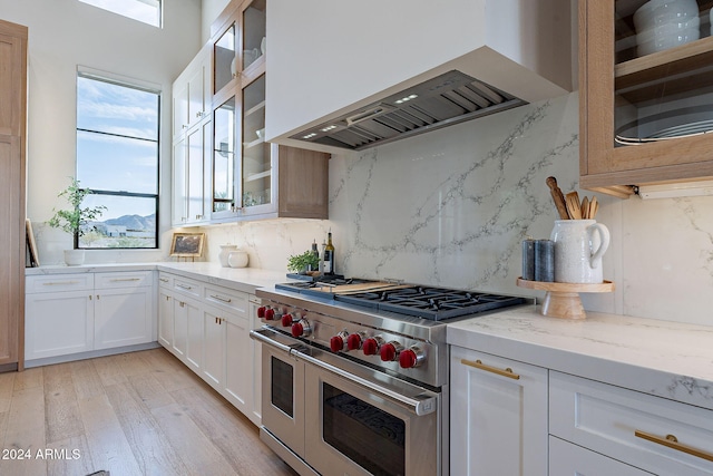 kitchen with wall chimney range hood, light stone countertops, double oven range, white cabinets, and light hardwood / wood-style floors