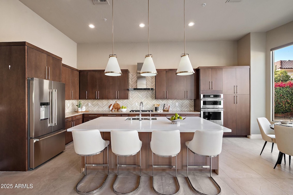 kitchen with hanging light fixtures, stainless steel appliances, wall chimney range hood, tasteful backsplash, and dark brown cabinets