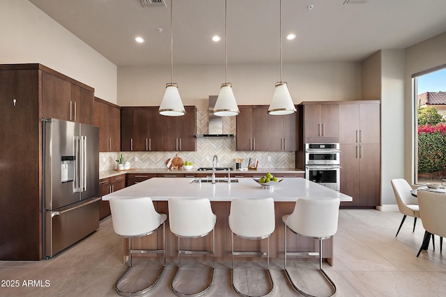 kitchen with hanging light fixtures, stainless steel appliances, wall chimney range hood, tasteful backsplash, and dark brown cabinets