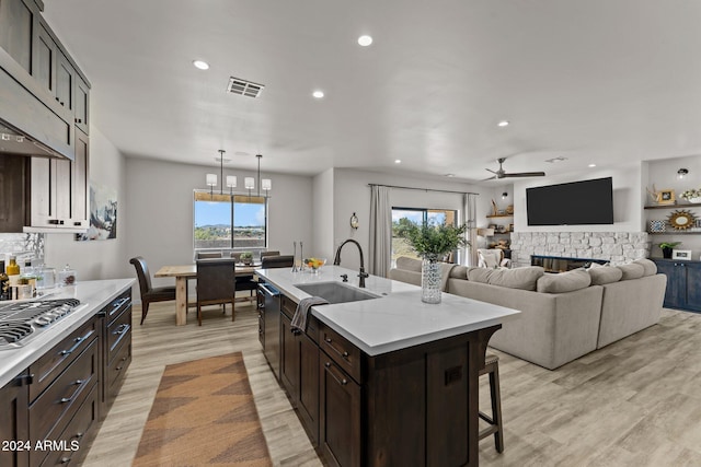 kitchen featuring sink, light wood-type flooring, a stone fireplace, a kitchen island with sink, and pendant lighting