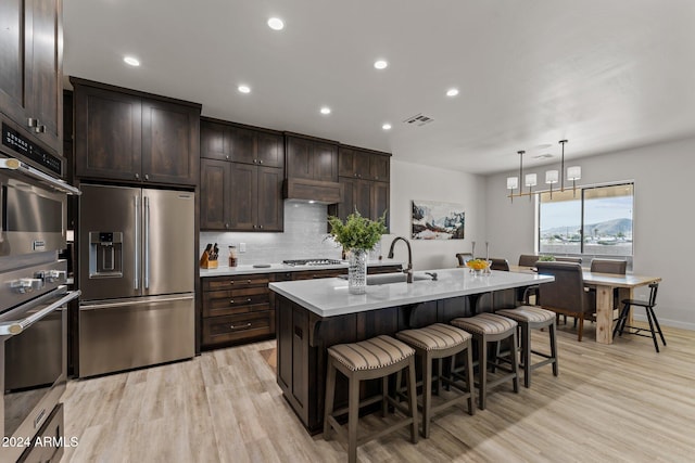 kitchen with a center island with sink, sink, appliances with stainless steel finishes, decorative light fixtures, and light hardwood / wood-style flooring