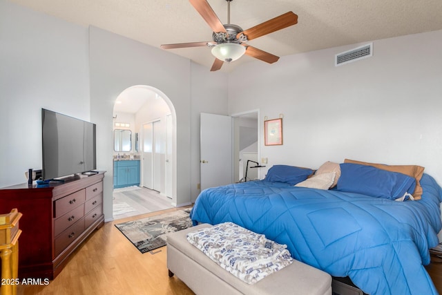 bedroom featuring visible vents, arched walkways, ensuite bathroom, a textured ceiling, and light wood-style floors