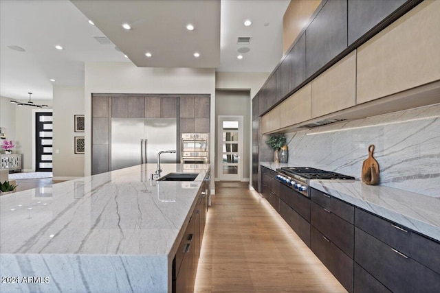 kitchen featuring a large island with sink, stainless steel appliances, sink, light stone countertops, and light wood-type flooring