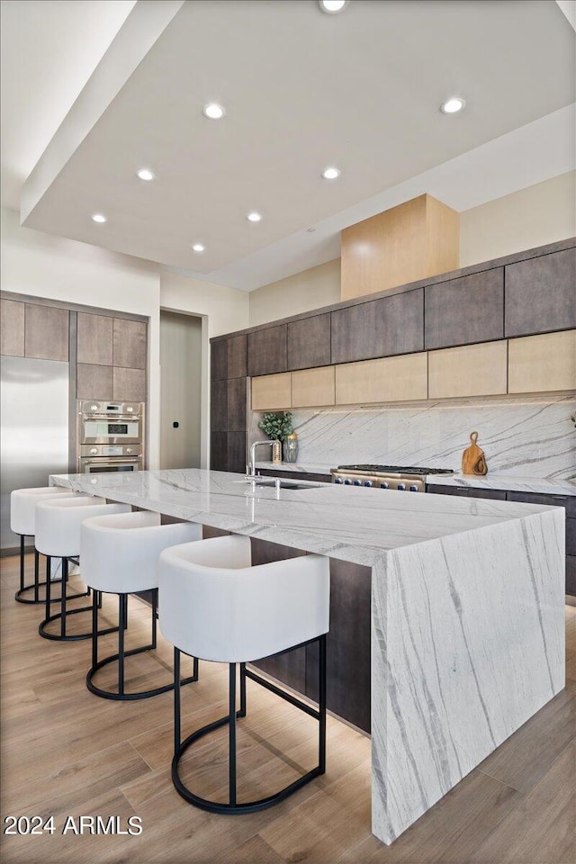 kitchen featuring a large island with sink, backsplash, light stone countertops, and light wood-type flooring