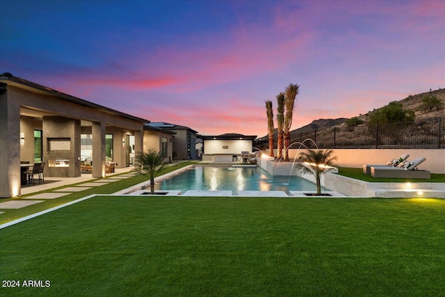 pool at dusk with a patio area, pool water feature, a lawn, and exterior kitchen