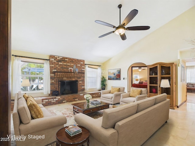 living room with ceiling fan, high vaulted ceiling, arched walkways, a brick fireplace, and light tile patterned floors