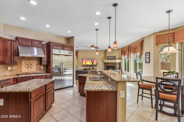 kitchen with a glass covered fireplace, an island with sink, stainless steel appliances, under cabinet range hood, and a sink