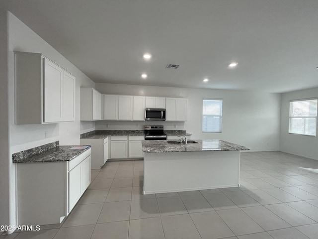 kitchen with white cabinets, dark stone counters, a center island with sink, light tile patterned flooring, and appliances with stainless steel finishes