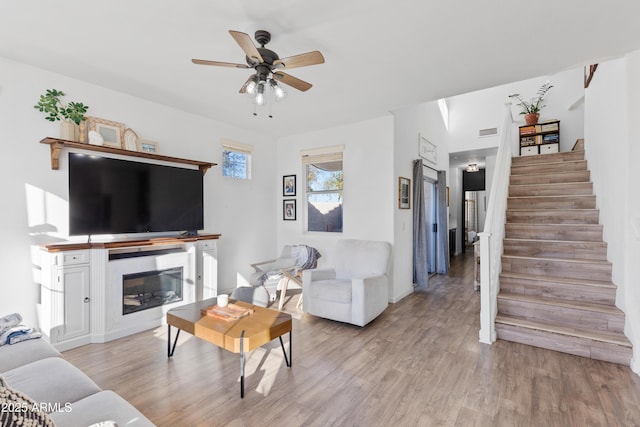 living area featuring a ceiling fan, visible vents, stairs, light wood-style floors, and a glass covered fireplace