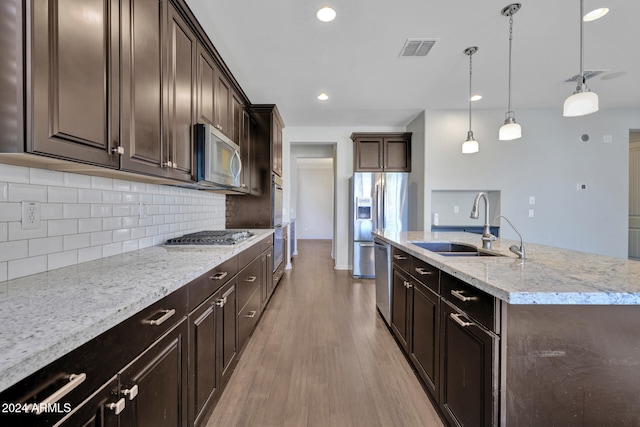 kitchen with appliances with stainless steel finishes, light wood-type flooring, light stone counters, a center island with sink, and hanging light fixtures