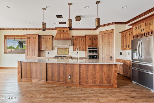 kitchen with appliances with stainless steel finishes, a sink, decorative backsplash, a large island with sink, and brown cabinetry