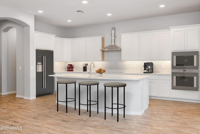 kitchen with an island with sink, stainless steel appliances, light hardwood / wood-style floors, and wall chimney range hood