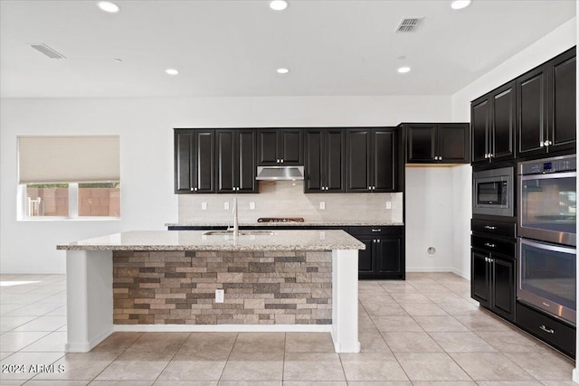 kitchen with light tile patterned flooring, sink, light stone counters, appliances with stainless steel finishes, and a kitchen island with sink